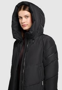 Svart vadderad jacka med en stor huva, dragkedja framtill och sidofickor. Har en mjuk, matt yta och sydda diagonala mönster.