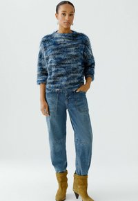 Blau gestreifter, flauschiger Pullover mit lockerer Passform, kombiniert mit hellblauen, weit geschnittenen Jeans. Getragen mit hellbraunen Stiefeletten und dezenten Creolen.