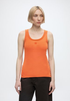 Mujer con cabello rubio corto, vestida con una camiseta sin mangas naranja con el logo "HUGO" y pantalones oscuros, de pie frente a un fondo blanco liso.