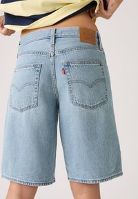 Άτομο που σηκώνει τη λαιμόκοψη του πουκαμίσου, φοράει ανοιχτό γαλάζιο denim σορτς Levi's με ορατές πίσω τσέπες σε ουδέτερο φόντο.