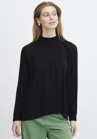 Haut noir à manches longues avec une texture côtelée et un col haut, présentant une coupe décontractée et un ourlet froncé, associé à un pantalon vert.