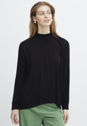 Black long-sleeve top with a ribbed texture and high neckline, featuring a relaxed fit and gathered hem, paired with green trousers.