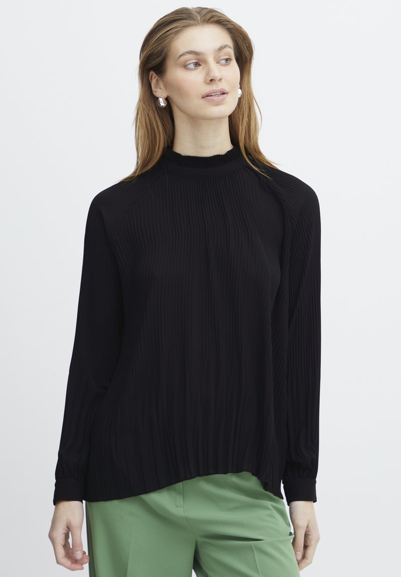 Haut noir à manches longues avec une texture côtelée et un col haut, présentant une coupe décontractée et un ourlet froncé, associé à un pantalon vert.