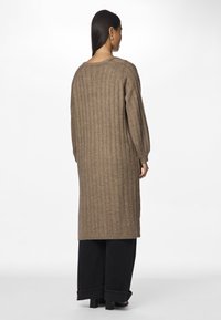 Pikk, pruun kudum cardigan, millel on vertikaalne sooniktekstuur, langetatud õlad ja mugav lõige. Kanda lahtiste mustade pükstega, millel on laiad pahkluupesad.