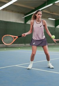 Kvinne i en lys lilla tanktopp og grå skort, som holder en oransje tenniskølle, klar til å slå en grønn tennisball på en blå tennisbane.