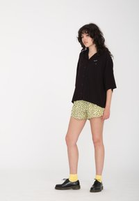 Chemise noire à manches courtes, assortie à un short jaune avec un motif abstrait noir, et des chaussures noires avec des chaussettes jaunes.