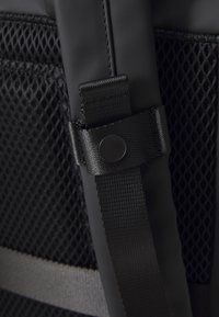 Black backpack detail showing textured mesh pocket, adjustable strap with a sleek buckle, and streamlined design for functional use.