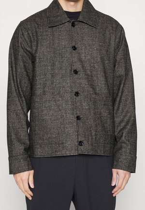 Dunkelgraue gemusterte Jacke mit Knöpfen und Kragen, über schwarzen Hosen getragen, dargestellt vom mittleren Oberkörper bis zum Hals, Hände entspannt an den Seiten.