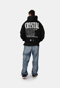 Felpa nera con cappuccio caratterizzata dalla scritta "CRYSTAL PARIS" in grande bianco, con un piccolo emblema della bandiera francese in basso, e jeans blu a vestibilità rilassata.