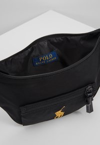 Svart tygpåse med en broderad hästlogotyp i guld, utrustad med dragkedjestängning och ett litet framfack med dragkedja. Inuti fodrad med svart material.