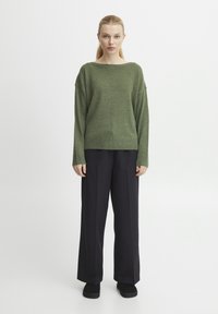 Grüner Strickpullover mit weitem Ausschnitt, kombiniert mit lockeren schwarzen Hosen und schwarzen Schuhen. Glatte Textur und entspannte Passform.