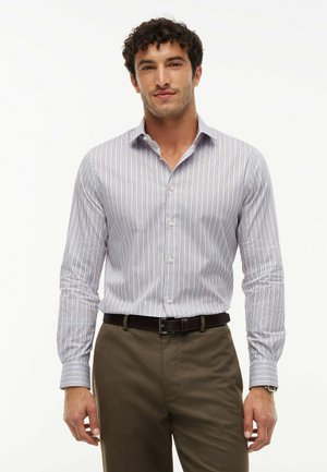 Homme portant une chemise à rayures verticales boutonnée, un pantalon marron et une ceinture sombre, se tenant devant un fond blanc uni.