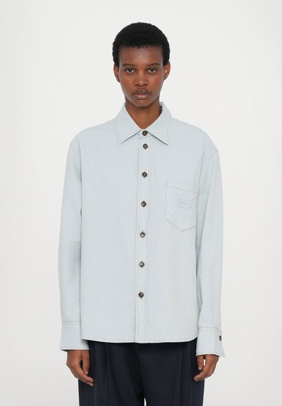 Chemise boutonnée bleu clair avec un col classique, poche avant et boutons marron. Coupe décontractée et manches longues avec des détails de couture subtils.