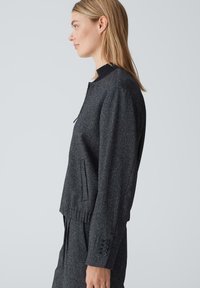 Graue, strukturierte Jacke mit hohem Kragen und gerippten Details. Besitzt seitliche Taschen und Knopfverzierungen an den Ärmeln.