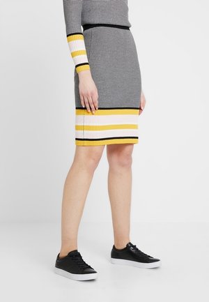 Vrouw draagt een aansluitende geribbelde gebreide rok en bijpassende top met zwarte, gele, witte en grijze horizontale strepen, gecombineerd met zwarte sneakers.
