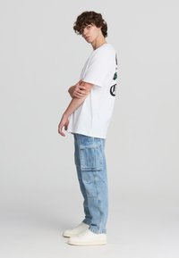 T-shirt bianca in cotone con stampa grafica sul retro, abbinata a jeans cargo azzurri chiari con tasche a toppa e sneakers bianche.