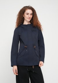 Jeune femme aux cheveux roux bouclés portant une veste bleu marine avec des poches avant et un pantalon noir, debout devant un fond blanc uni.