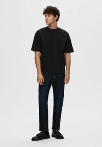 Tricou negru cu mânecă scurtă și guler rotund, asortat cu blugi de culoare albastră închisă. Modelul stă pe un fundal neutru, afișând o ținută casual.