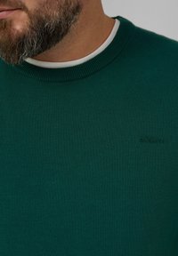 Groene trui met een ribbelboord aan de ronde hals, gemaakt van een gestructureerde brei-stof. Heeft een subtiel logo-detail aan de voorkant.