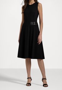 Svart ärmlös klänning med figursydd midja och utsvängd kjol. Har ett texturerat svart bälte med dubbel spänne. Slät tygtextur.