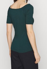 Teal kortärmad topp med en bred båthals och figursydd siluett, tillverkad av mjukt material, med en slät textur och rundad avslutning.