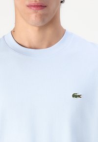 Tricou din bumbac de culoare albastru deschis, cu decolteu rotund și un mic logo verde în formă de crocodil pe pieptul stâng. Textură netedă, croială casual.