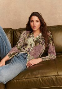 Bloemenpatroon overhemd in lila en groentinten, gecombineerd met lichtblauwe jeans, op een donkergroene leren bank. Natuurlijke textuur.