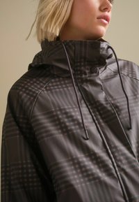 Chaqueta gris con patrón y cuello alto, cuenta con capucha, cordones ajustables y botones de presión, mostrando un diseño a cuadros y textura suave.