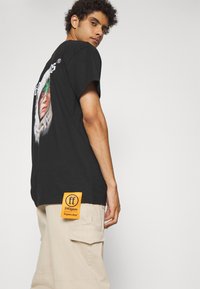 Sort bomulds t-shirt med et grafisk print og et orange label med tekst. Modellen bærer beige cargo bukser med sidelommer.