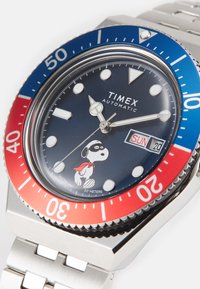Zegarek automatyczny Timex ze stalową bransoletą. Bezel w kolorze niebiesko-czerwonym, granatowa tarcza, białe indeksy godzinowe oraz detal postaci Snoopy. Wodoodporny do 50 m.