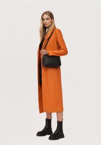 Oranžový dlouhý kabát z hladkého materiálu, spárovaný s černou prošívanou crossbody taškou a černými kotníkovými botami.