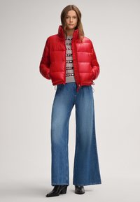 Czerwona puchowa kurtka z wysokim kołnierzem, zamkiem błyskawicznym i elastycznymi sznurkami; zestawiona z niebieskimi dżinsami o szerokich nogawkach i czarnymi botkami na obcasie.