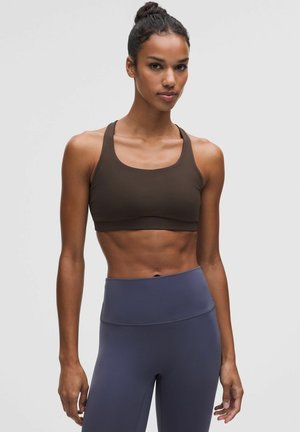 Fit vrouw met donker haar in een knot draagt een donkerbruine sportbeha en hooggetailleerde donkerblauwe leggings tegen een effen grijze achtergrond.