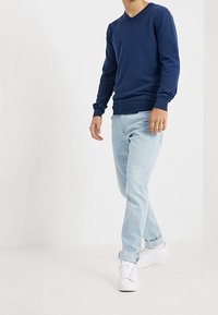 Persona che indossa una maglietta a maniche lunghe blu navy, jeans chiari arrotolati e scarpe da ginnastica bianche, che cammina in uno spazio bianco semplice.