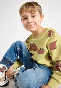 Groene sweatshirt met dierenprints, gecombineerd met blauwe spijkerbroek. Schoenen zijn beige met zwarte accenten. Sokken hebben een berenontwerp.