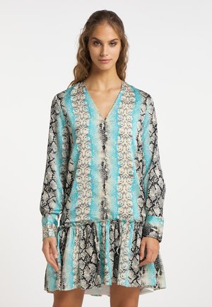 Vrouw draagt een mini-jurk met lange mouwen en V-hals, met blauw, zwart en beige slangenprint en geplooide zoom, staand tegen een witte achtergrond.