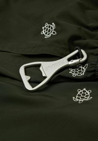 Décapsuleur mousqueton argenté de marque Iuman reposant sur un tissu foncé avec des motifs brodés de tortues marines blanches.