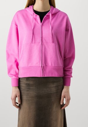 Femme portant un sweat à capuche rose vif zippé avec cordons et une jupe marron foncé, debout devant un fond clair uni.