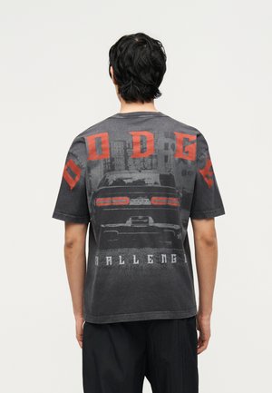 Mężczyzna widziany od tyłu, ubrany w ciemnoszarą koszulkę z czerwonymi literami „DODGE” oraz grafiką przedstawiającą tylny widok samochodu i budynków miejskich.