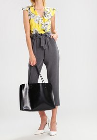Donna che indossa un top giallo a fiori con volant, pantaloni grigi a vita alta con tasca a sacchetto, tacchi bianchi, mentre tiene una grande borsa a tracolla nera al suo fianco.
