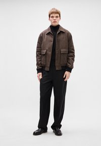 Casaco de mistura de lã castanho com colarinho, dois bolsos frontais, sobre uma camisola de gola alta preta, combinado com calças e sapatos pretos.