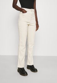 Jeans de veludo cotelê creme, com corte flare, cintura alta e bolsos laterais. Usados com botas pretas com atacadores, apresentando uma textura suave e nervuras verticais.