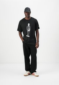 Μαύρο μπλουζάκι που διαθέτει γραφιστικό σχέδιο ενός φιγούρας, σε συνδυασμό με μαύρο παντελόνι και λευκά παπούτσια με κόκκινες ρίγες. Το καπέλο είναι επίσης μαύρο.