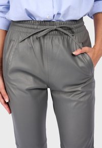 Šedé kožené kalhoty s elastickým pasem, stahovací šňůrkou a bočními kapsami. Povrch má hladkou, měkkou strukturu.