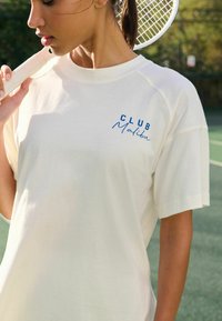Krämfärgad t-shirt med kort ärm och en avslappnad passform. Funktioner med blå broderad text "CLUB Malibu" på vänster bröst. Mjuk textur.