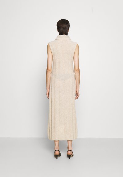 Vestido bege sem mangas, com gola alta, comprimento longo, textura suave e padrão vertical subtil, combinado com sapatos de salto pretos.