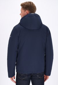 Mornarsko modra zip-up jakna z velikim kapuco, z gladko teksturo in preprostim dizajnom, oblečena čez temne kavbojk.
