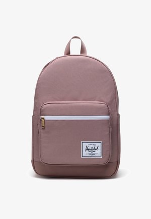 Sac à dos en tissu rose de forme arrondie, avec une poche zippée à l'avant, une étiquette tissée et une poignée supérieure. Texture lisse avec un design minimaliste.