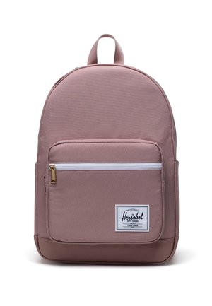 Sac à dos en tissu rose de forme arrondie, avec une poche zippée à l'avant, une étiquette tissée et une poignée supérieure. Texture lisse avec un design minimaliste.