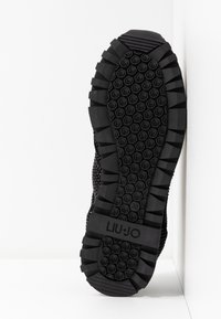 Semelle de chaussure de sport noire avec caoutchouc texturé, présentant un motif en zigzag et des grips circulaires, logo en relief et indication de taille.